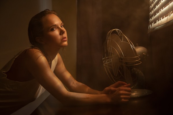 Young woman in a white tank top suffering from the heat sitting in front of an old fan with louver lighting by Artmood Visualz